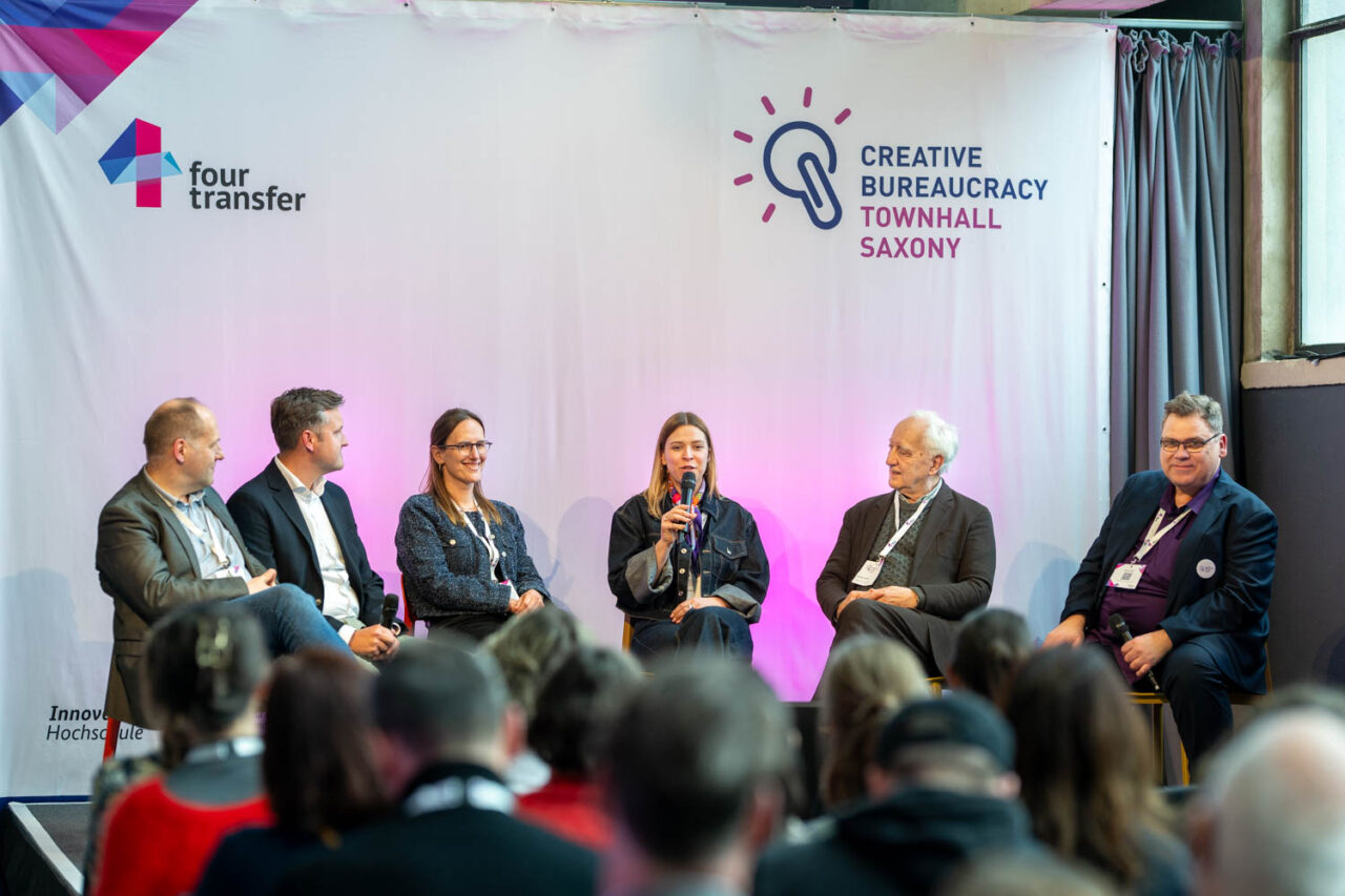 Das Bild zeigt eine Aufnahme des Panels "Seismograph Townhall - Bisherige Erkenntnisse und Creative Bureaucracy Formate der Zukunft. Zu sehen sind unter anderem Prof. Dr. Oliver Jokisch von der HSF Mei0en, Creative Bureaucracy Festival Gründer Charles Landry, die ehemalige CBF Festivaldirektorin Johanna Sieben, Townhall Saxony Projektleiter Christof Schreckenberg und Erik Friemel von der Sächsischen Aufbaubank (SAB) sowie Caroline Hoffmann von der Kreativbehörde Regensburg.